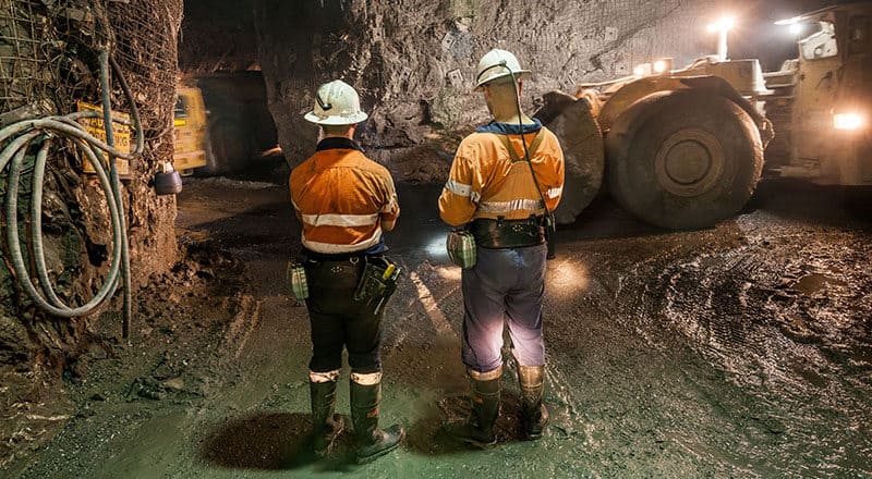 Two workers in a well-lit mine with yellow mining machines.