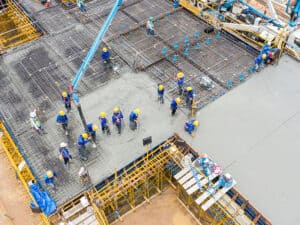 Areal view of a construction site where workers in blue uniforms and yellow hard hats are using a concrete pump.