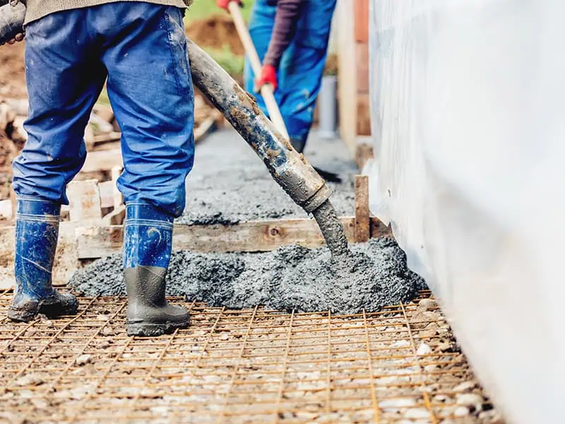 Construction worker in blue rubber boots and pants pumping concrete into a building foundation.