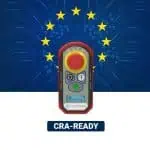 Bezdrátové dálkové ovládání Cattron s tlačítkem nouzového zastavení na pozadí modré desky s plošnými spoji, na které jsou vyobrazeny hvězdy EU a nápis „CRA-READY“.