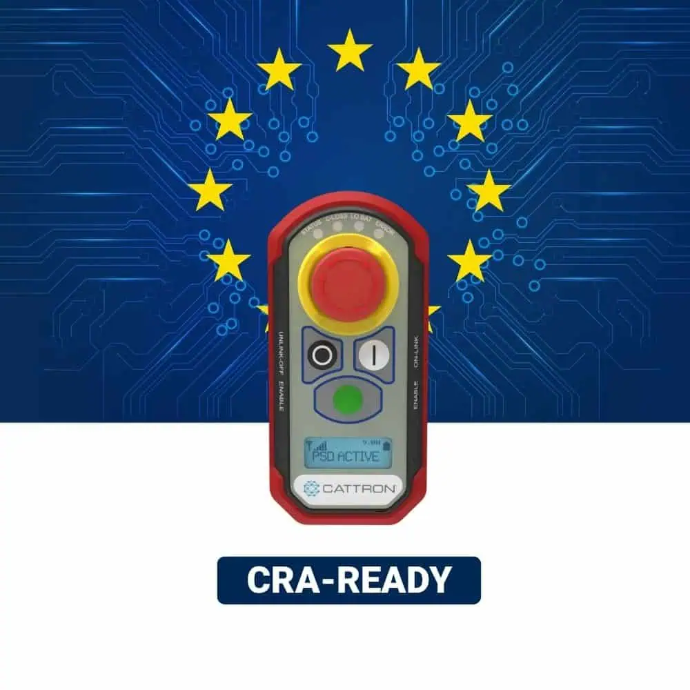 Bezdrátové dálkové ovládání Cattron s tlačítkem nouzového zastavení na pozadí modré desky s plošnými spoji, na které jsou vyobrazeny hvězdy EU a nápis „CRA-READY“.